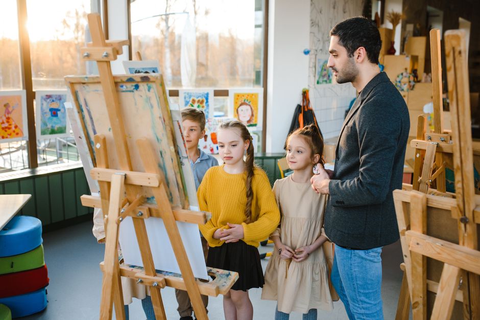 Children painting on easels under a teacher's guidance, illustrating how AI shifts classroom work.