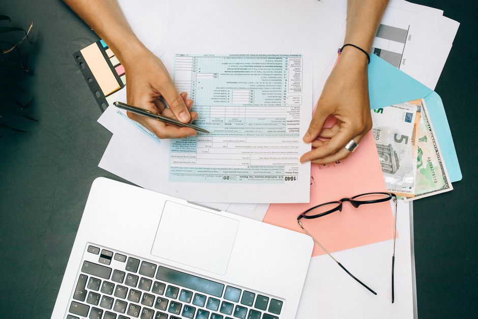 Stock photo of hands writing on tax documents with laptop, glasses, and currency on desk, used to illustrate this story on CBIC’s 26 October GST Guidance Date Has Passed.