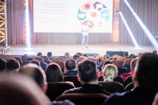 An audience watches a presentation with stage lights and a speaker, illustrating the Goa Conclave discussions on university models and AI.