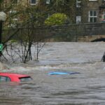 Three cars partially submerged in a flooded river near a stone bridge, illustrating storm disruption in the UK.
