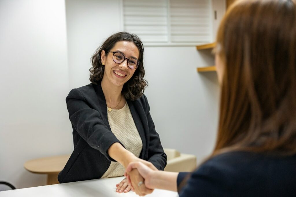 Two women shake hands across a table, illustrating discussion of reduced junior technical hiring amid expanding AI use.