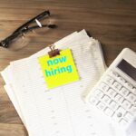 Stock photo of Calculator, glasses, and papers with 'now hiring' note, used to illustrate this story on How to Set and Defend Your First Salary Offer.