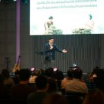 Stock photo of A man speaking into a microphone on stage before an audience in a hall, used to illustrate this story on Europe Adds to Business Regulatory Burden, Speaker Says.