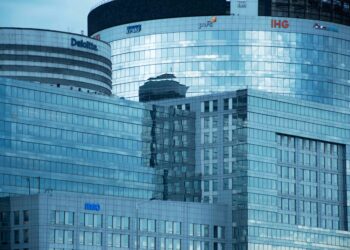 Glass skyscrapers reflecting the sky, showcasing brand logos under a calm sky.